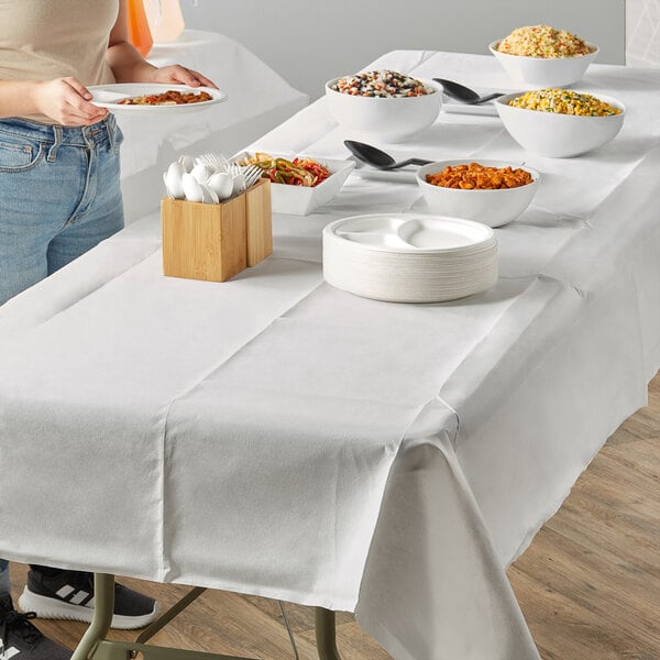 A woman standing next to a table with a Hoffmaster white table cover filled with plates of food.