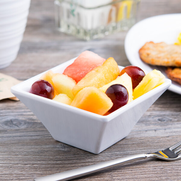 A white squared melamine bowl filled with fruit on a table.