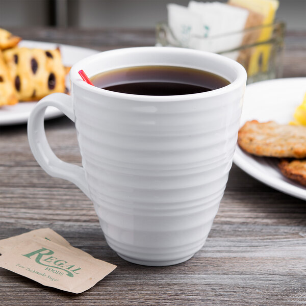 A white Elite Global Solutions melamine mug filled with coffee on a table next to a plate of food.