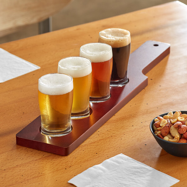 An Acopa mahogany flight paddle with pub tasting glasses of beer and a bowl of mixed nuts on a table.