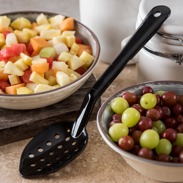 A bowl of fruit salad with a Carlisle black perforated serving spoon next to a bowl of grapes.