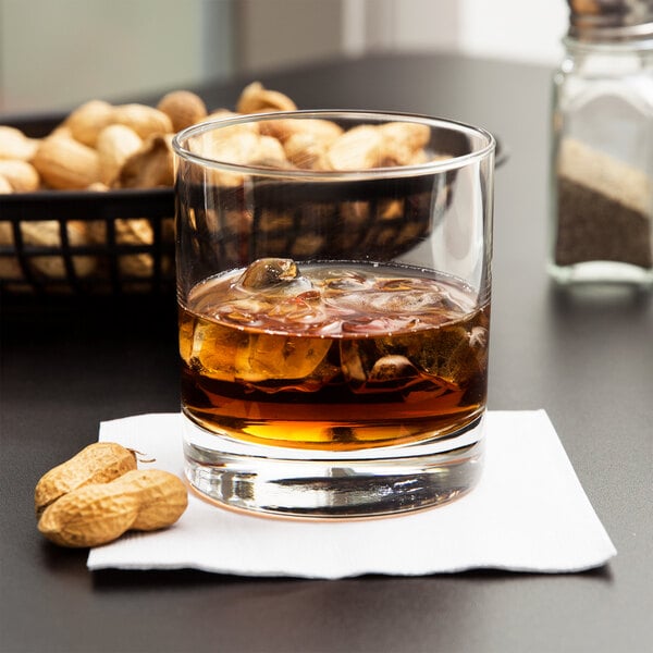 A Libbey rocks glass filled with ice and whiskey on a table with a basket of peanuts.
