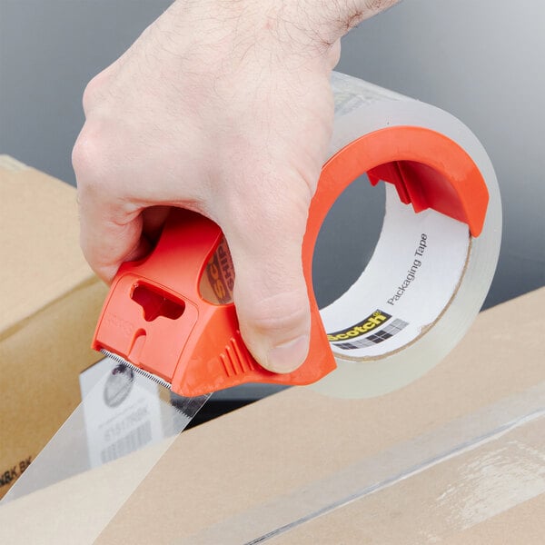 A hand using a 3M Scotch heavy-duty tape dispenser to seal a box.