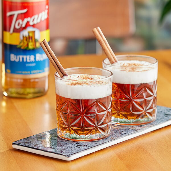 Two glasses of Torani Butter Rum flavoring syrup with brown liquid and cinnamon sticks.