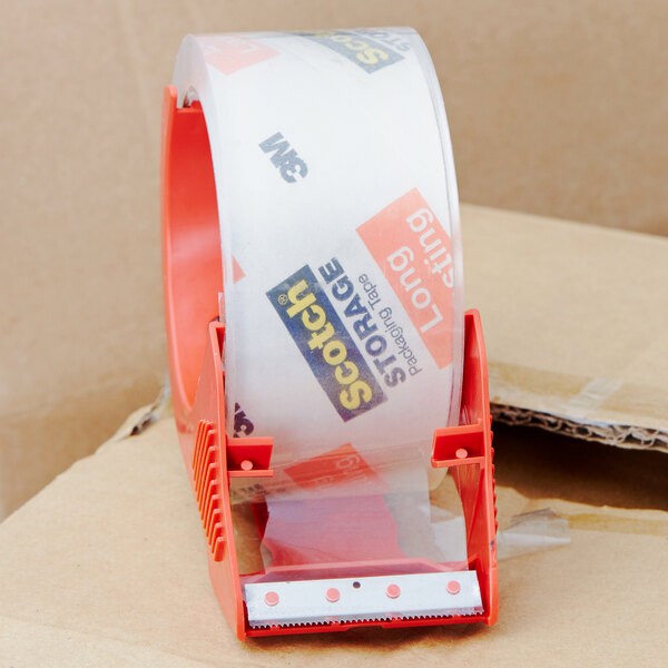 A 3M Scotch clear tape dispenser sitting on a cardboard box with a roll of tape on top.