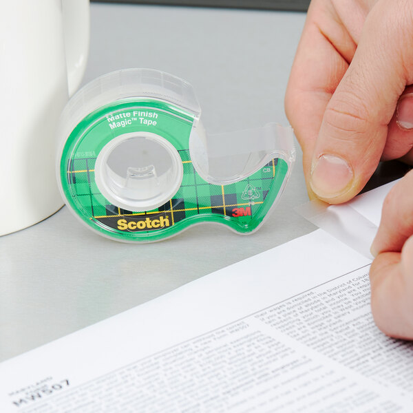 A hand using a green and white 3M Scotch tape dispenser to write on paper with transparent 3M Scotch Magic tape.
