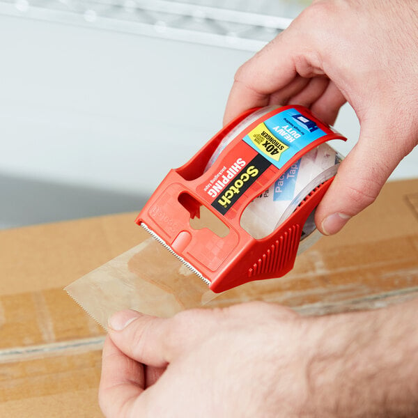 A hand using a 3M Scotch heavy-duty red tape dispenser to seal a box.