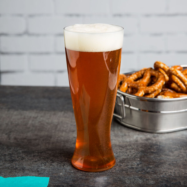 A WNA Comet clear plastic pilsner glass filled with beer on a table next to a bowl of pretzels.