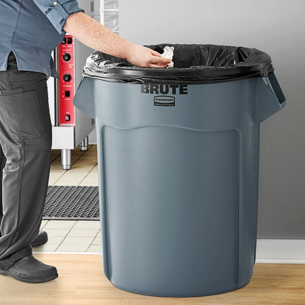 A man standing next to a Rubbermaid Brute 55 gallon gray round trash can.