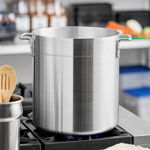 A Choice standard weight aluminum stock pot on a stove with a wooden spoon with holes on top.