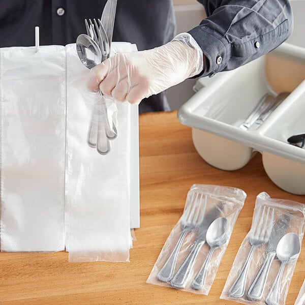 A gloved hand holding a plastic bag of silverware with a fork and spoon inside.