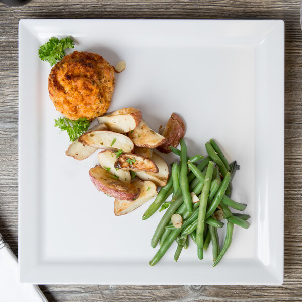 A white square Elite Global Solutions melamine plate with a meat patty, green beans, and a potato wedge on a wood table.