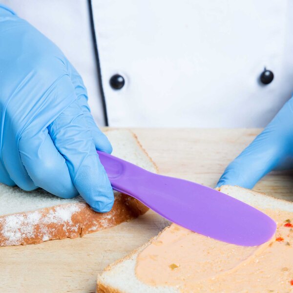 A person in blue gloves using a purple HS Inc. sandwich spreader to spread a spread on a piece of bread.