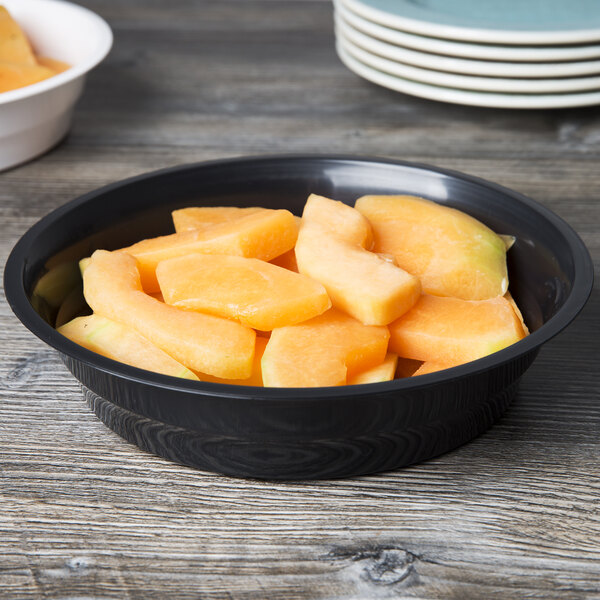 A black Fineline low profile plastic serving bowl filled with sliced fruit on a table.