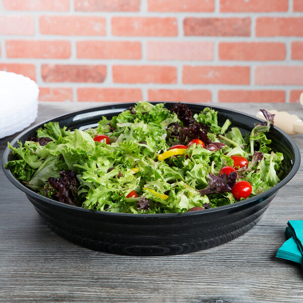 A black Fineline low profile plastic bowl filled with salad on a table.
