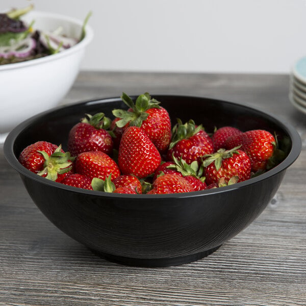 A black Fineline high profile plastic catering bowl filled with strawberries on a table with a bowl of salad.