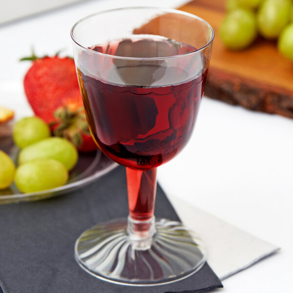 A close up of a Fineline plastic wine goblet filled with red liquid on a table next to green grapes.
