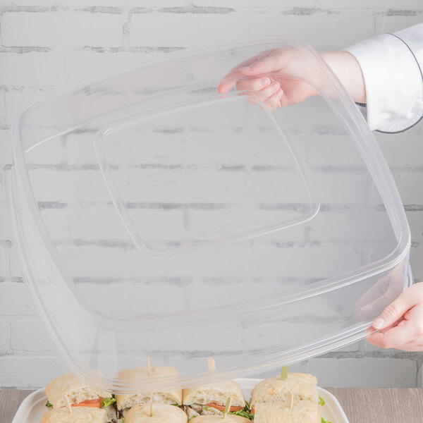 A hand holding a clear plastic container with a lid over a tray of sandwiches.