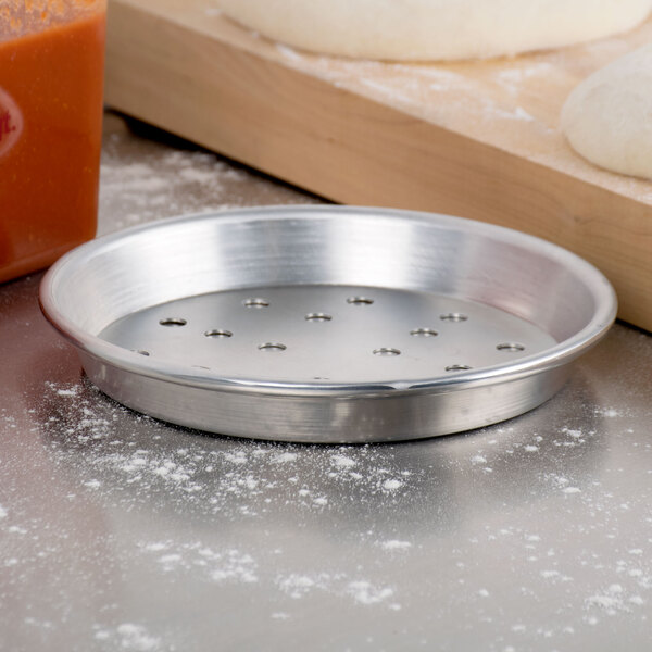 An American Metalcraft silver aluminum pizza pan with holes next to dough on a wooden cutting board.