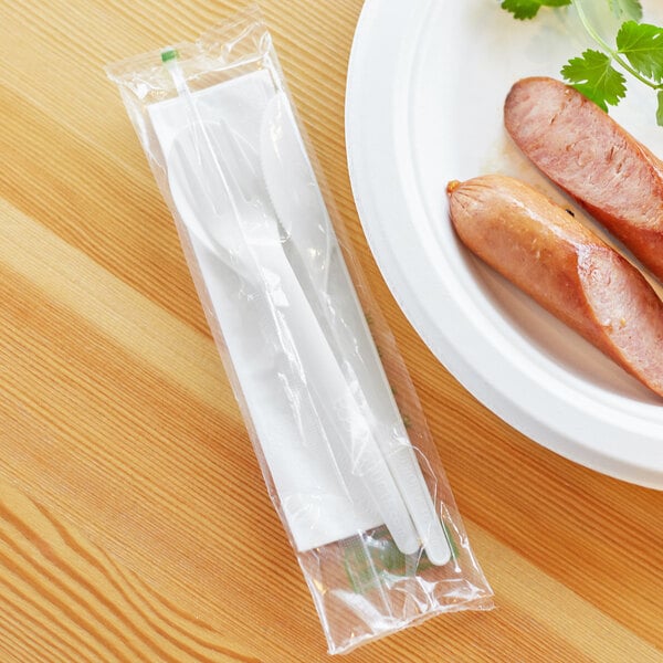 A plate of sausages with a fork and knife on it next to a plastic bag of white compostable utensils.
