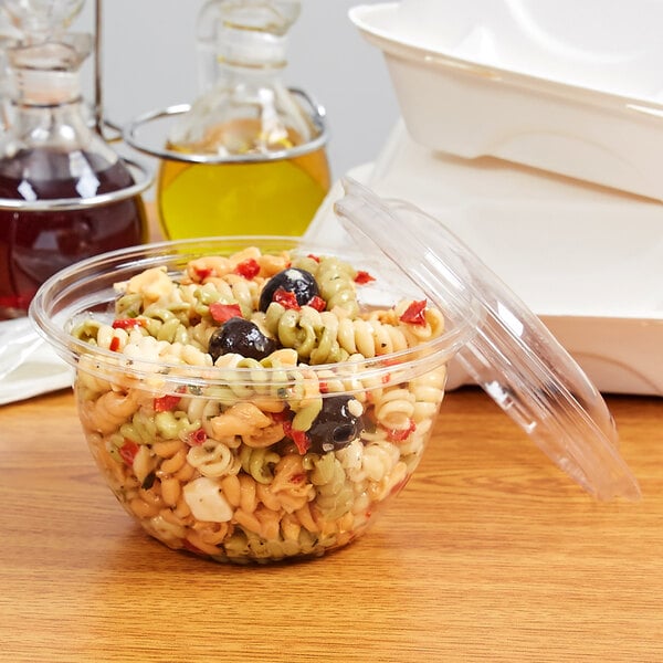 A clear plastic Eco-Products salad bowl with pasta, olives, and cheese on a table.