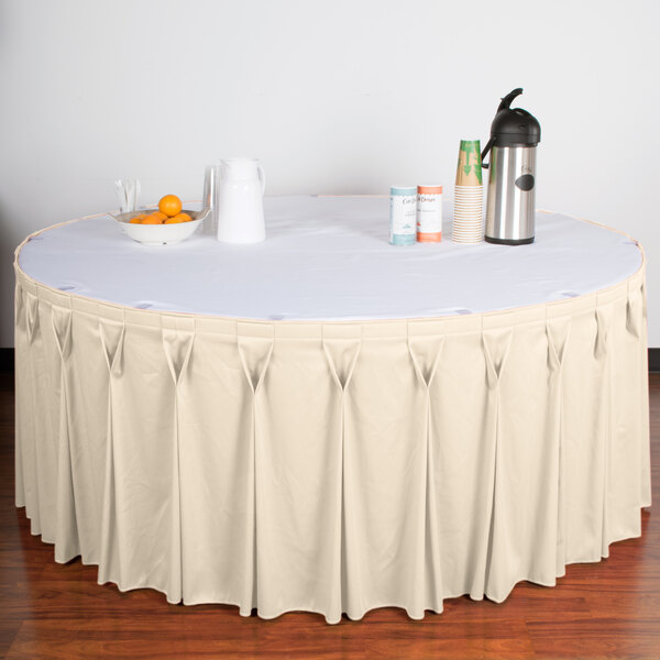 A table with a Snap Drape Wyndham bone bow tie pleat table skirt on a white table with a bowl of oranges and a thermos.