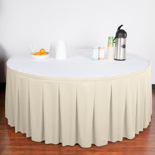 A table with a Snap Drape bone continuous pleat table skirt on a white table with a bowl of oranges and a thermos.