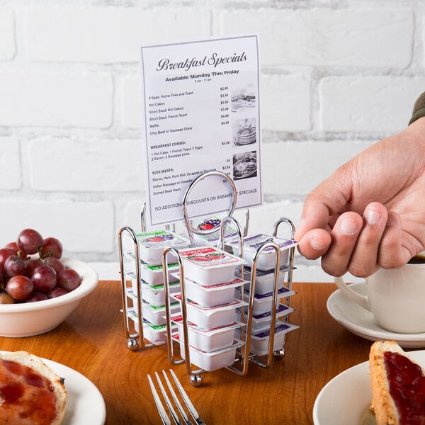 A hand holding a small container of jelly on a Tablecraft chrome plated rack on a table with breakfast.
