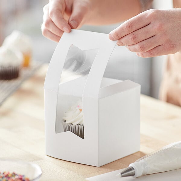 A person opening a Baker's Mark white window cupcake box to reveal a cupcake inside.