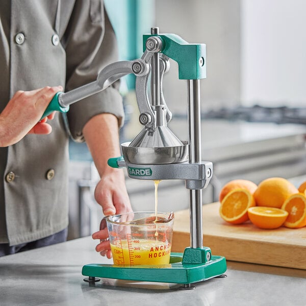 A person using the Garde manual citrus juicer to make orange juice with a measuring cup full of orange juice and a cut orange nearby.