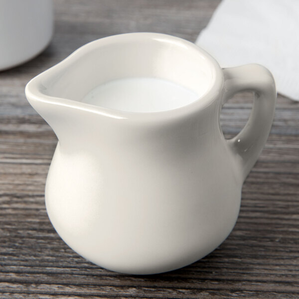 A white Tuxton china creamer on a table with a pitcher of milk.