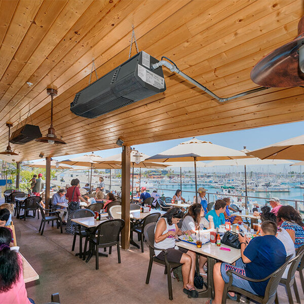 A group of people sitting at tables outside under a white umbrella with a view of the ocean using a Schwank natural gas black patio heater.