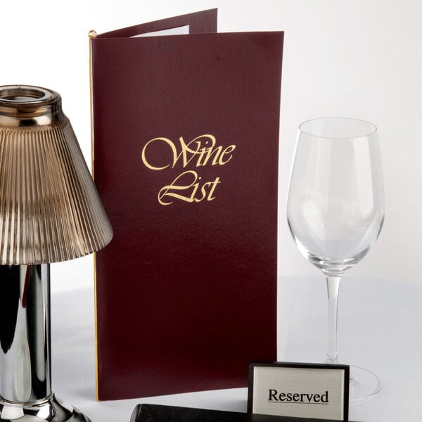 A burgundy Menu Solutions wine list cover on a table with a wine glass and a wine list.