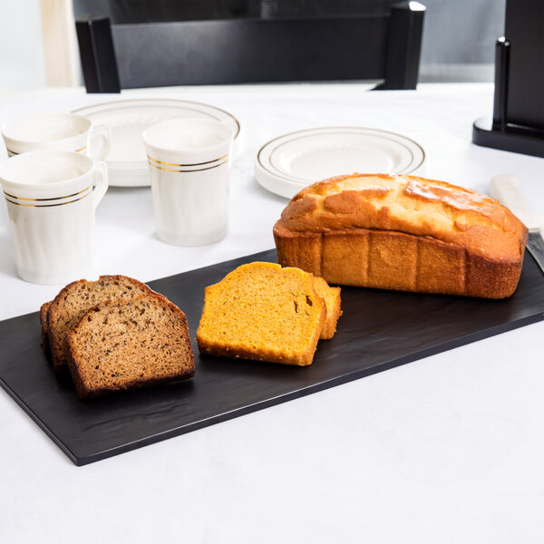 An American Metalcraft black faux slate melamine tray with a loaf of bread and a plate of muffins on a table.
