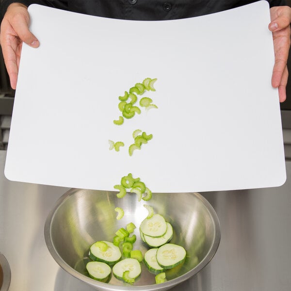 A person using a white Tablecraft flexible cutting board to cut cucumbers over a bowl.