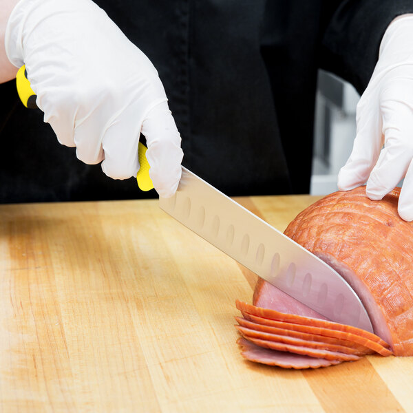 A hand wearing a white glove uses a Mercer Culinary Millennia Colors Santoku Knife with a yellow handle to cut meat.