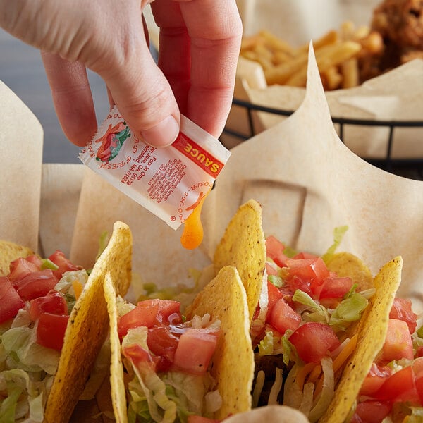 A person pouring Hot Sauce from a small packet onto a taco shell filled with tomatoes and lettuce.