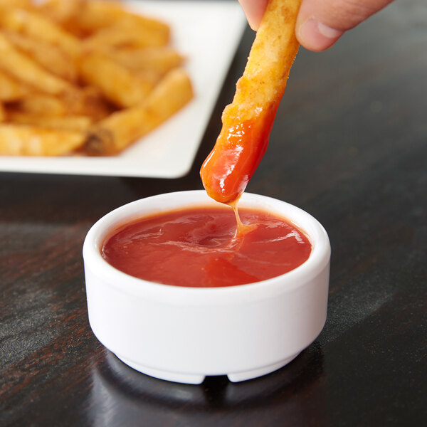 A person using a Carlisle white ramekin to dip a french fry into ketchup.