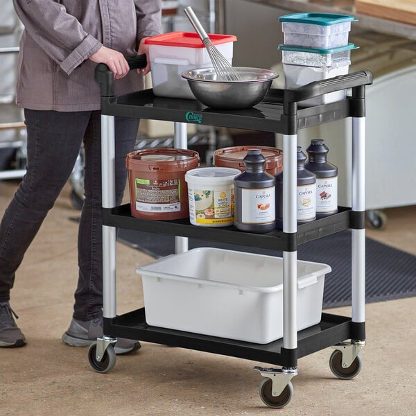 A woman pushing a Choice black utility cart with three shelves and white containers on it.