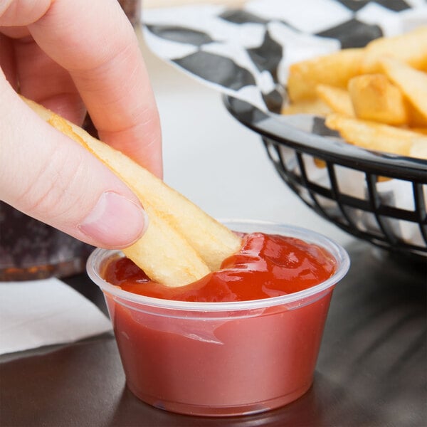 A hand holding a french fry and dipping it into a clear plastic container of ketchup.