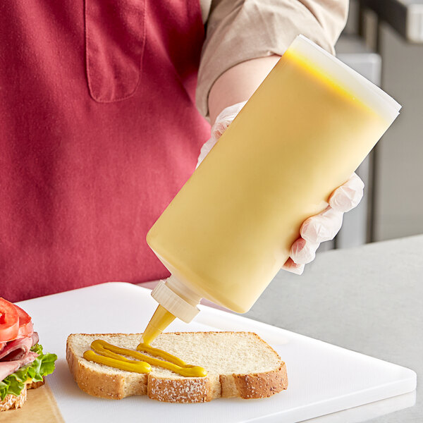 A person using a Prince Castle reusable squeeze bottle to pour mustard on a sandwich.