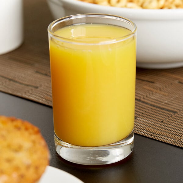 A Libbey side water/tasting glass filled with orange juice next to a bowl of cereal.
