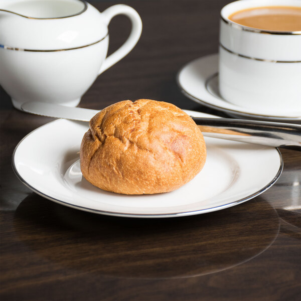 A 10 Strawberry Street Silver Line porcelain bread plate with a bread roll on it next to a cup of coffee.