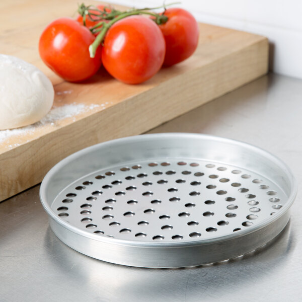 An American Metalcraft tin-plated steel pizza pan with holes on it next to tomatoes on a cutting board.
