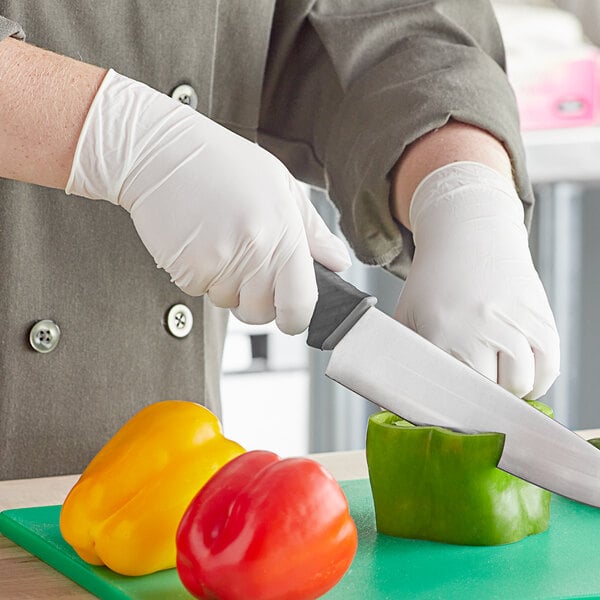 A person in a white coat cutting green bell peppers with a knife using Noble Products disposable gloves.