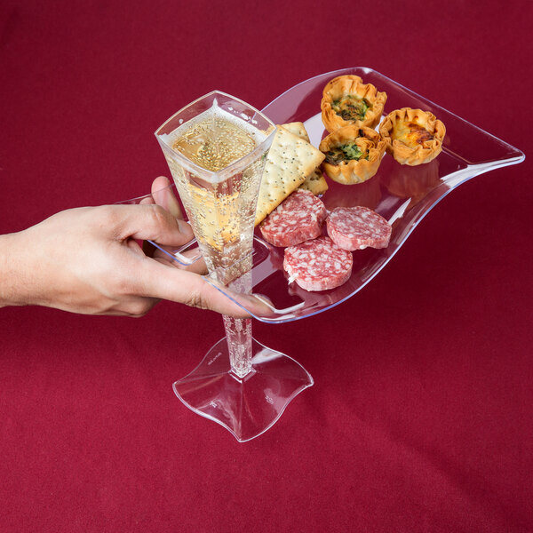 A hand holding a glass of champagne and a clear plastic cocktail plate with food on it.