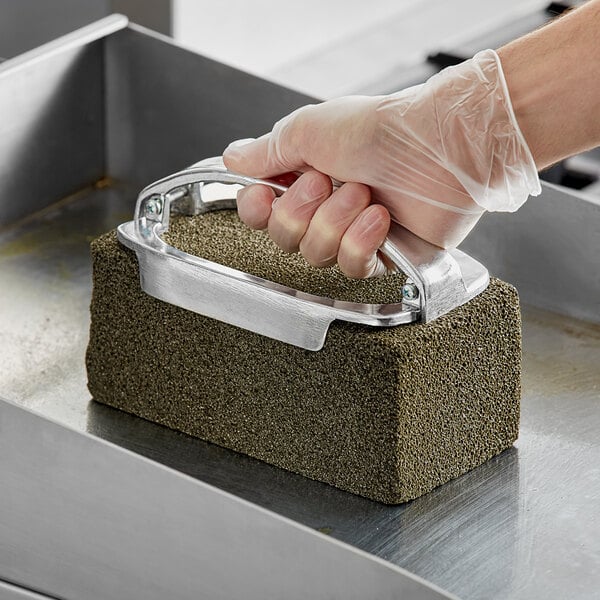 A person wearing a plastic glove uses a Scrubble grill brick to clean a metal surface.