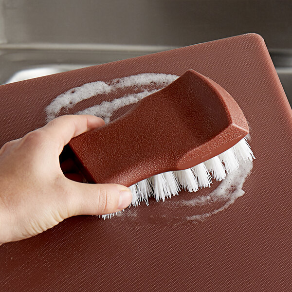 A hand using a brown Thunder Group cutting board brush to clean a red cutting board.