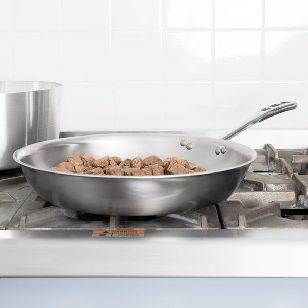 A Vollrath stainless steel fry pan with meat cooking on a stove.