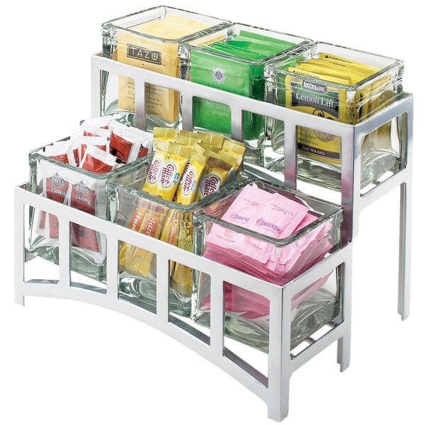 A Cal-Mil silver steel two tier display holding glass containers of various flavored tea packets.